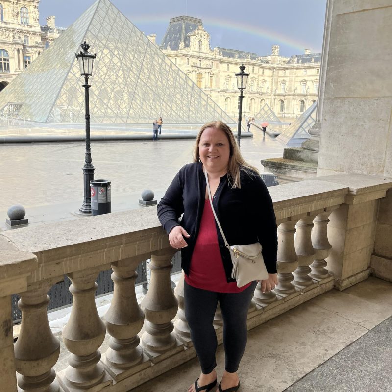 Andrea standing outside the Louvre with the pyramid and a rainbow in the background.